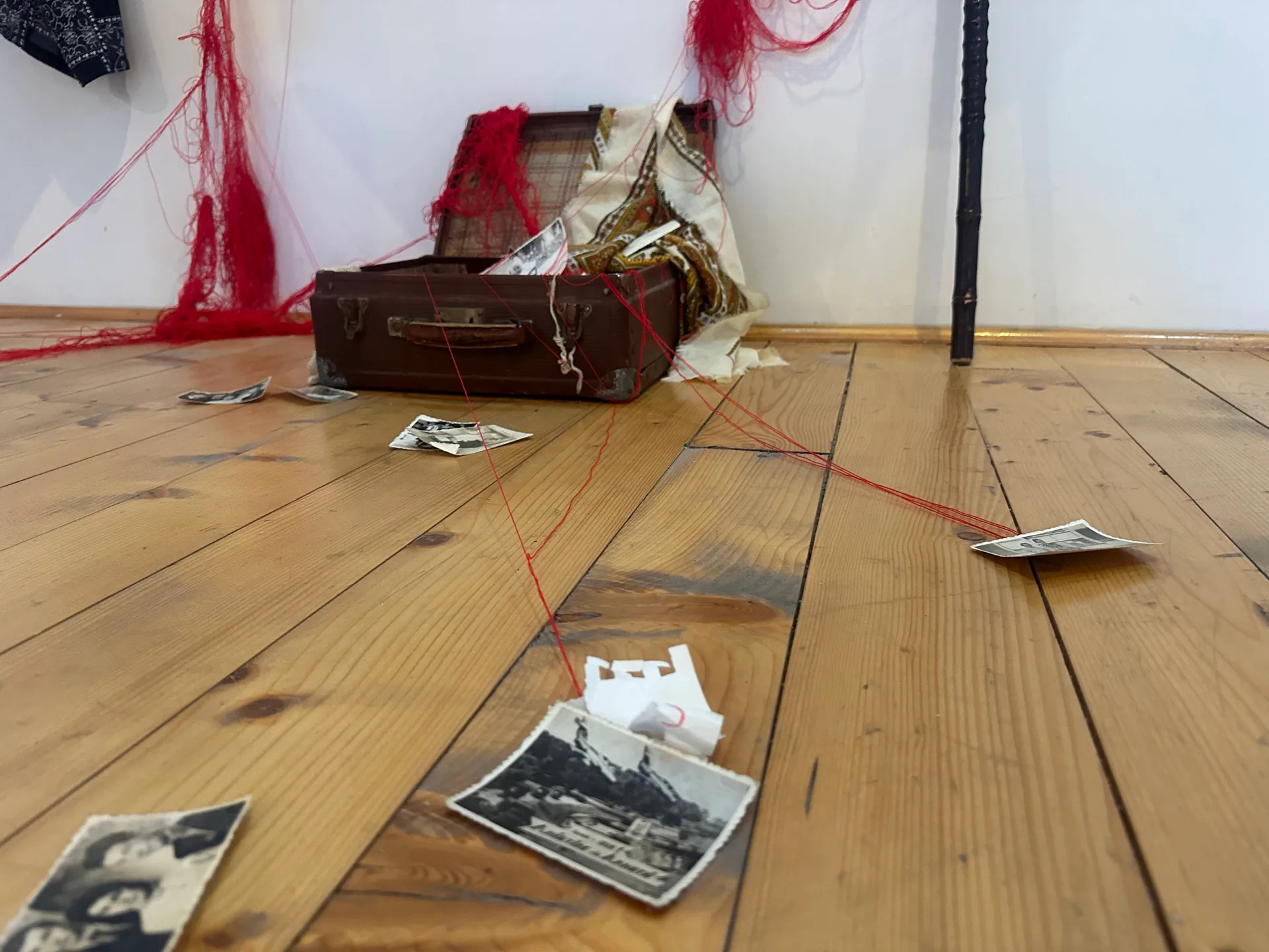 Lower half of the installation showing the hanging robe, red wool thread, open suitcase, walking stick, and slippers on the wooden gallery floor